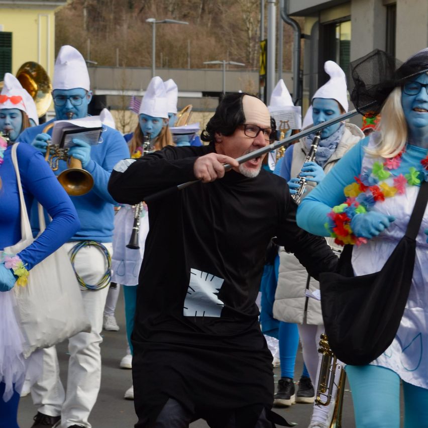 Eine Gruppe von Menschen in blauen und weißen Kostümen und Hüten zieht in einem Umzug, wobei ein Mann in einem schwarzen Kostüm einen Stab hält.