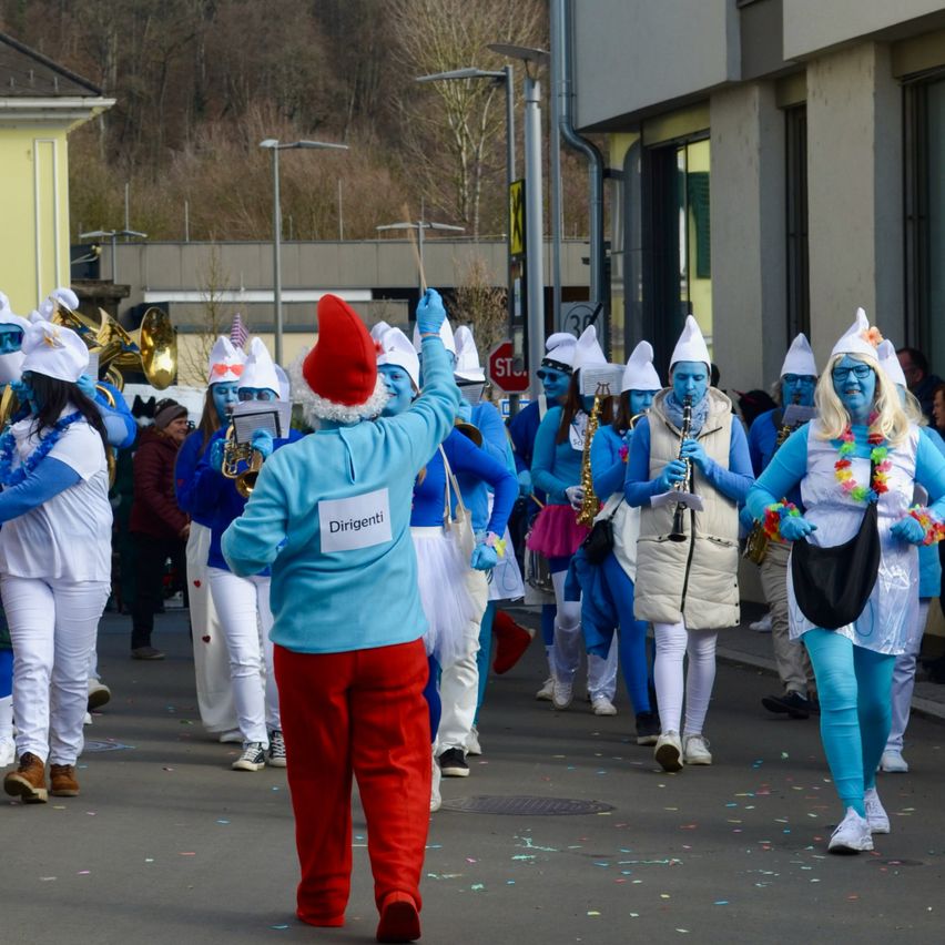 Eine Gruppe von Menschen, die in Blau gekleidet sind, marschiert in einer Parade und hält Musikinstrumente. Eine Person trägt einen roten Hut und rote Hosen, während andere in Weiß sind. Gebäude und Bäume sind im Hintergrund zu sehen.