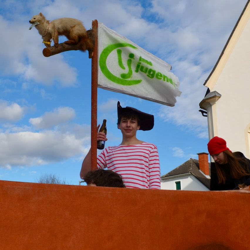 Ein junger Mann in einem gestreiften Shirt und Hut hält eine Flasche, während er neben einem Eichhörnchen steht, das ein Stück Papier hält. Eine weiße Flagge mit grünem Text steht auf einem Pfahl. Eine Frau mit einer roten Mütze beobachtet in der Nähe.