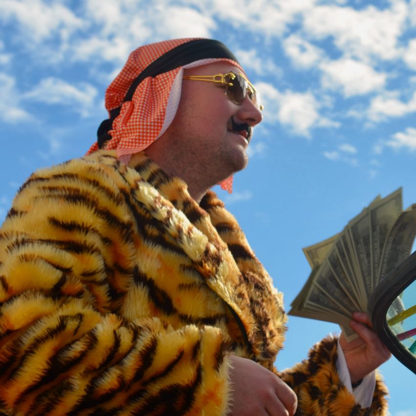 Ein Mann in einem Tigerkostüm, mit Kopftuch und Sonnenbrille, hält einen Haufen Geld. Der Himmel ist blau mit einigen Wolken.
