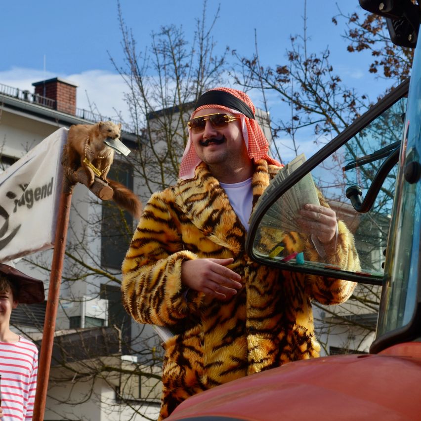 Ein Mann in einem Tigerkostüm steht neben einem Fahrzeug mit einem Eichhörnchen auf einem Schild mit der Aufschrift Jugend. Eine weitere Person in einem gestreiften Shirt steht in der Nähe.