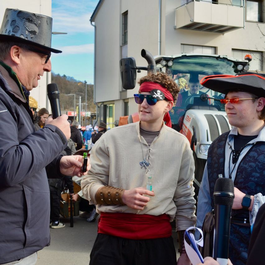 Drei Männer stehen vor einem Gebäude, einer hält ein Mikrofon. Der Mann in der Mitte in Piratenkostüm hält eine grüne Flasche. Der Mann rechts trägt einen Piratenhut und eine Brille. Ein Traktor ist hinter ihnen geparkt.