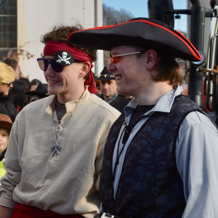 Zwei Männer in Piratenkostümen stehen nebeneinander und lächeln. Sie tragen Piratenhüte, Bandanas und Sonnenbrillen. Dahinter steht eine Menge Menschen in Piratenkostümen.