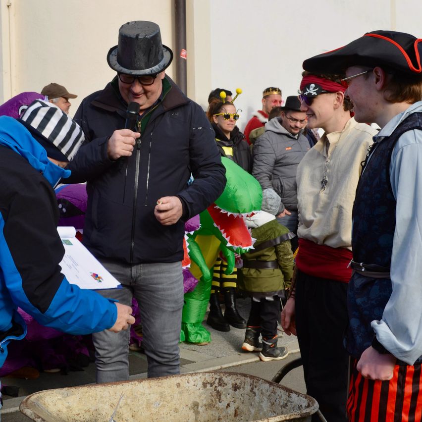Ein Mann in einem schwarzen Zylinder spricht in ein Mikrofon, während andere in Kostümen zusehen. Ein Mann in einem Piratenhut steht rechts von ihm. Ein Schubkarren steht in der Nähe.
