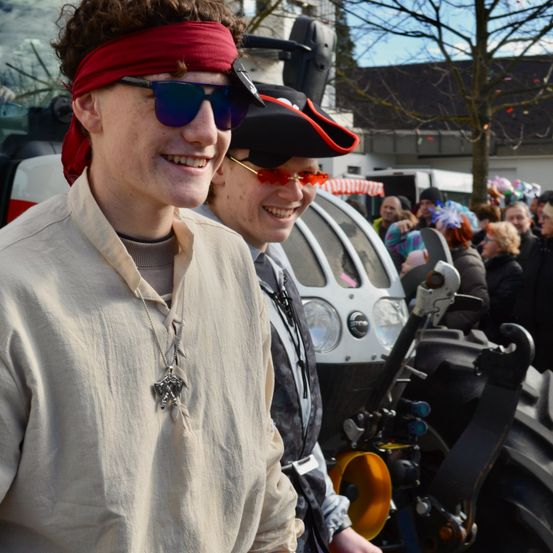 Zwei Menschen in mittelalterlichen Kostümen stehen vor einem Traktor bei einer Veranstaltung im Freien, mit einer Menschenmenge im Hintergrund.