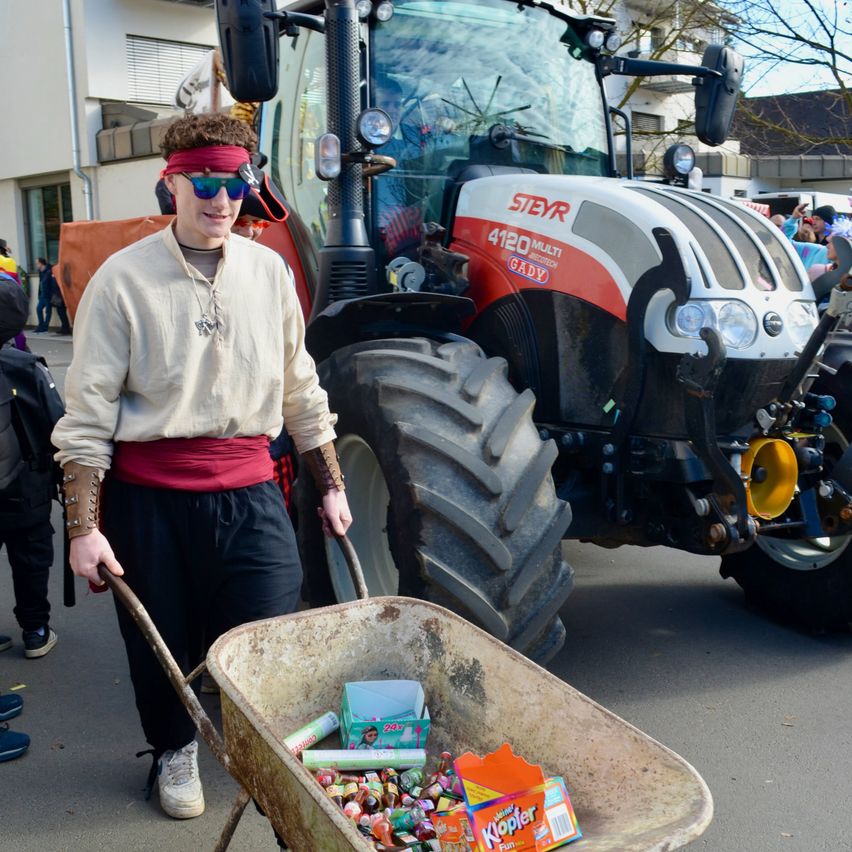 Eine Person in einem Kostüm schiebt einen mit Süßigkeiten gefüllten Schubkarren vor einem Traktor.