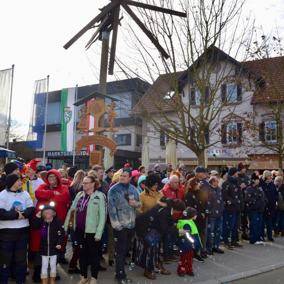 Eine große Menschenmenge versammelt sich vor einem Gebäude, wahrscheinlich für eine festliche Veranstaltung. Viele von ihnen tragen Hüte und Brillen. Einige von ihnen halten eine Seifenblasen-Pistole und erzeugen Seifenblasen. Ein hölzerner Pfahl mit einem Schild und einem Wappen steht in der Mitte der Menge. Ein Baum steht vor dem Gebäude und eine Fahne ist auf der linken Seite.