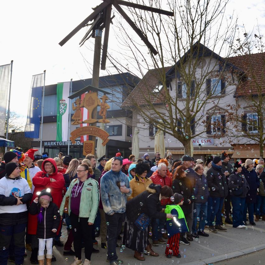 Eine große Menschenmenge versammelt sich vor einem Gebäude, wahrscheinlich für eine festliche Veranstaltung. Viele von ihnen tragen Hüte und Brillen. Einige von ihnen halten eine Seifenblasen-Pistole und erzeugen Seifenblasen. Ein hölzerner Pfahl mit einem Schild und einem Wappen steht in der Mitte der Menge. Ein Baum steht vor dem Gebäude und eine Fahne ist auf der linken Seite.