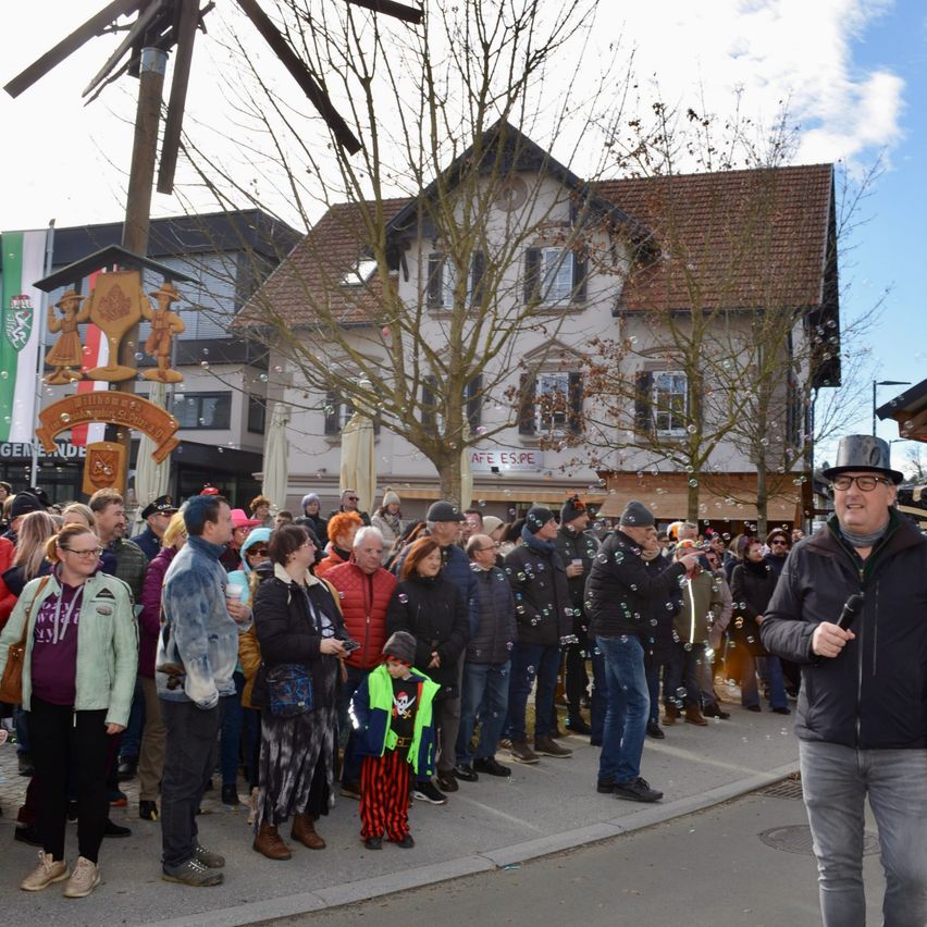 Eine Menschenmenge versammelt sich vor einem Haus mit einer Fahne und Bäumen. Einige Menschen tragen Winterkleidung, und ein Mann in einem Piratenkostüm bläst Seifenblasen.