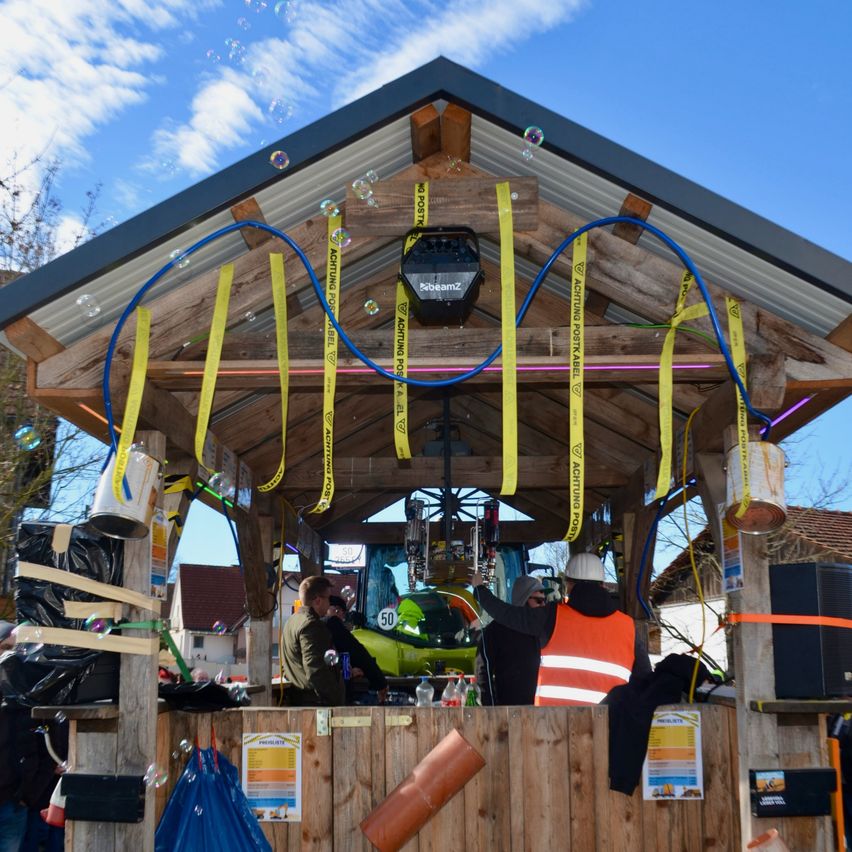 Eine Holzstruktur mit einem Metalldach, dekoriert mit gelben Bändern und blauen Schläuchen. Eine Person in einer orangefarbenen Weste steht im Inneren, mit einem schwarzen Lautsprecher und einem Traktor im Blickfeld. Andere Personen sind anwesend, zusammen mit verschiedenen Gegenständen wie Flaschen und einem blauen Beutel. Die Umgebung scheint sich im Freien unter einem klaren blauen Himmel zu befinden.