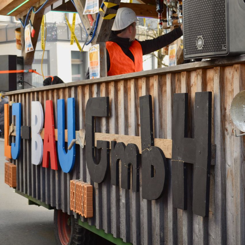 Ein Mann in einer orangefarbenen Weste steht hinter einem Tresen mit den Buchstaben J-BAU GmbH, die prominent darauf zu sehen sind. Er bereitet etwas vor, möglicherweise für ein Festival.