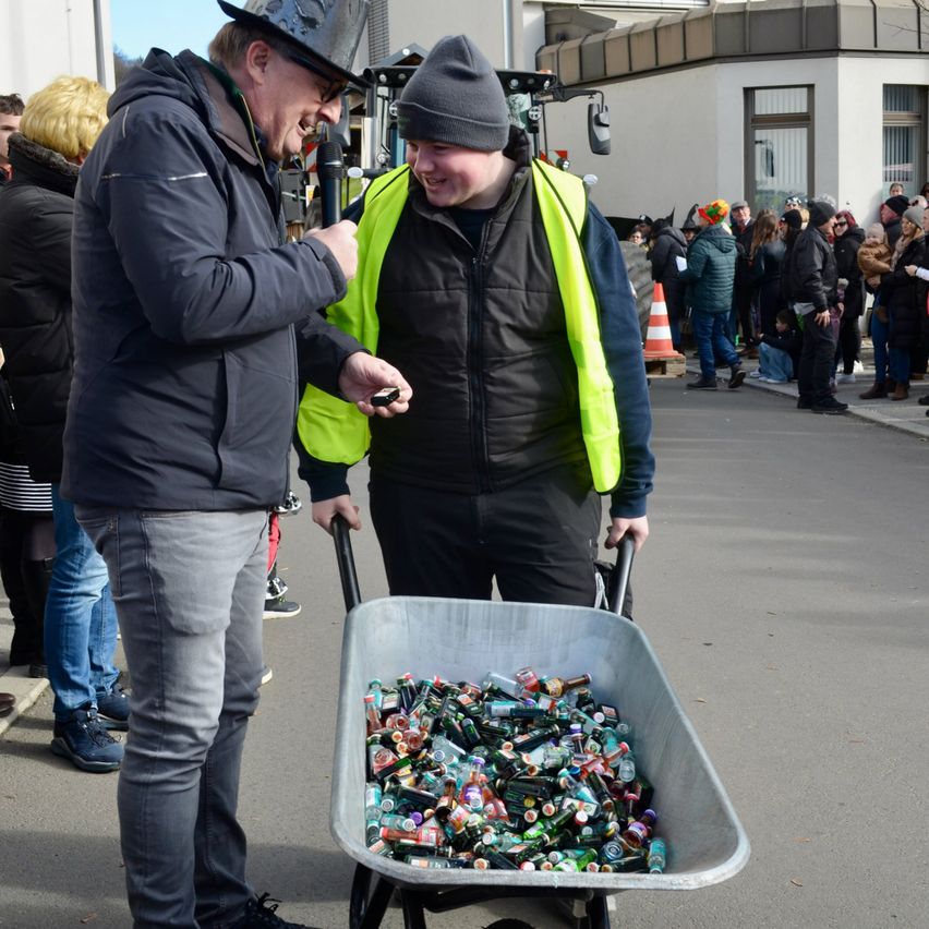 Ein Mann mit Hut schiebt einen mit Limonadendosen gefüllten Schubkarren, während ein Mann neben ihm lacht. Sie sind von einer Menge Menschen umgeben.