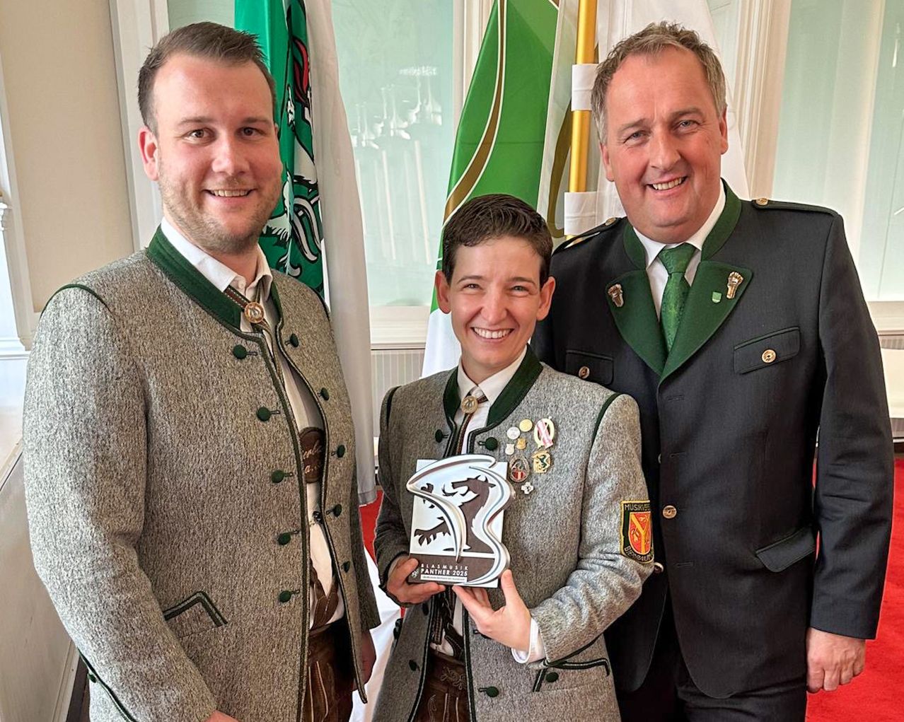 Drei Männer stehen vor Fahnen, einer hält eine Trophäe. Der Mann in der Mitte trägt eine Medaille.