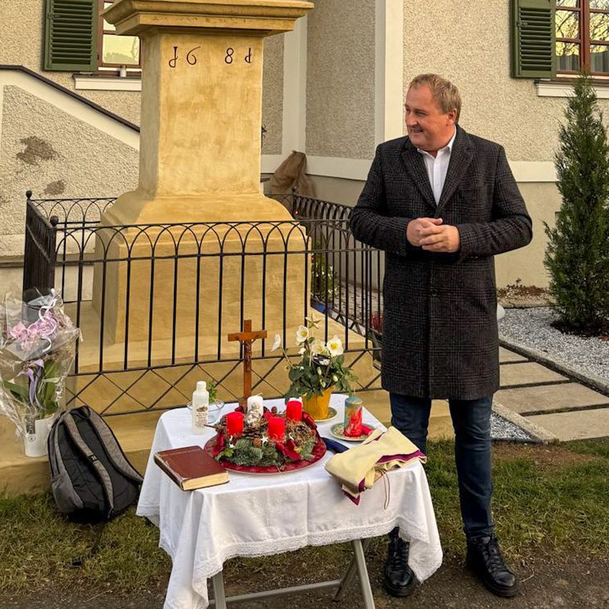 Ein Mann steht vor einem Denkmal mit einem Tisch, auf dem Blumen, Kerzen und ein Buch stehen. Das Denkmal ist mit einem Kreuz und einer kleinen Topfpflanze geschmückt.