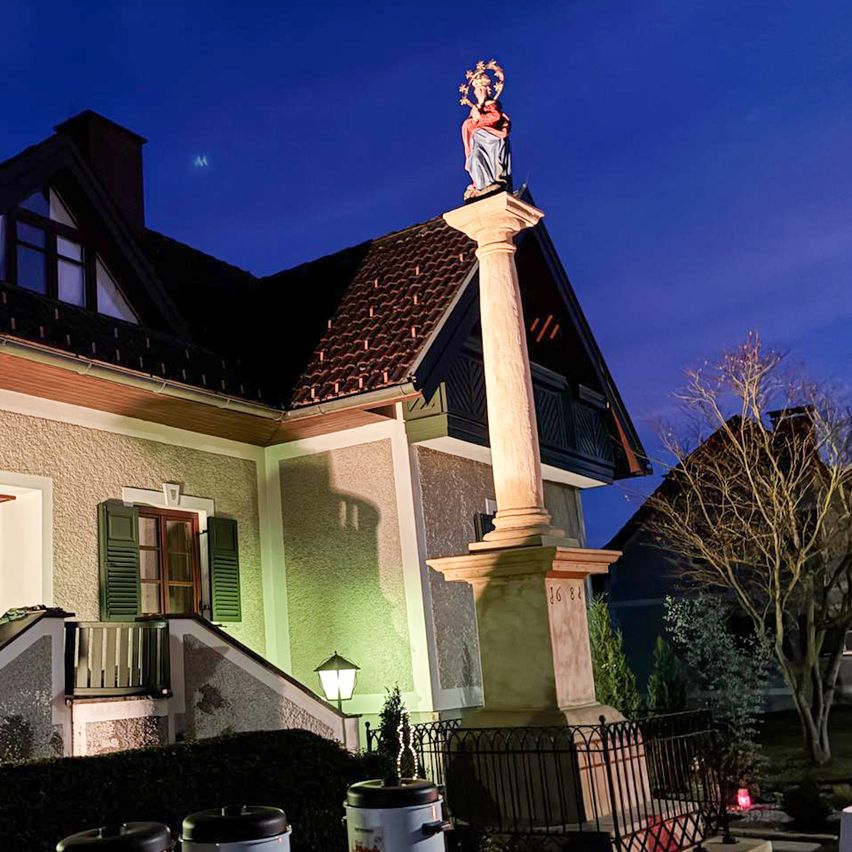 Eine Statue einer Frau mit einem Kranz und einem Kind steht auf einer Säule vor einem Haus. Das Haus hat grüne Fensterläden und ist nachts beleuchtet.