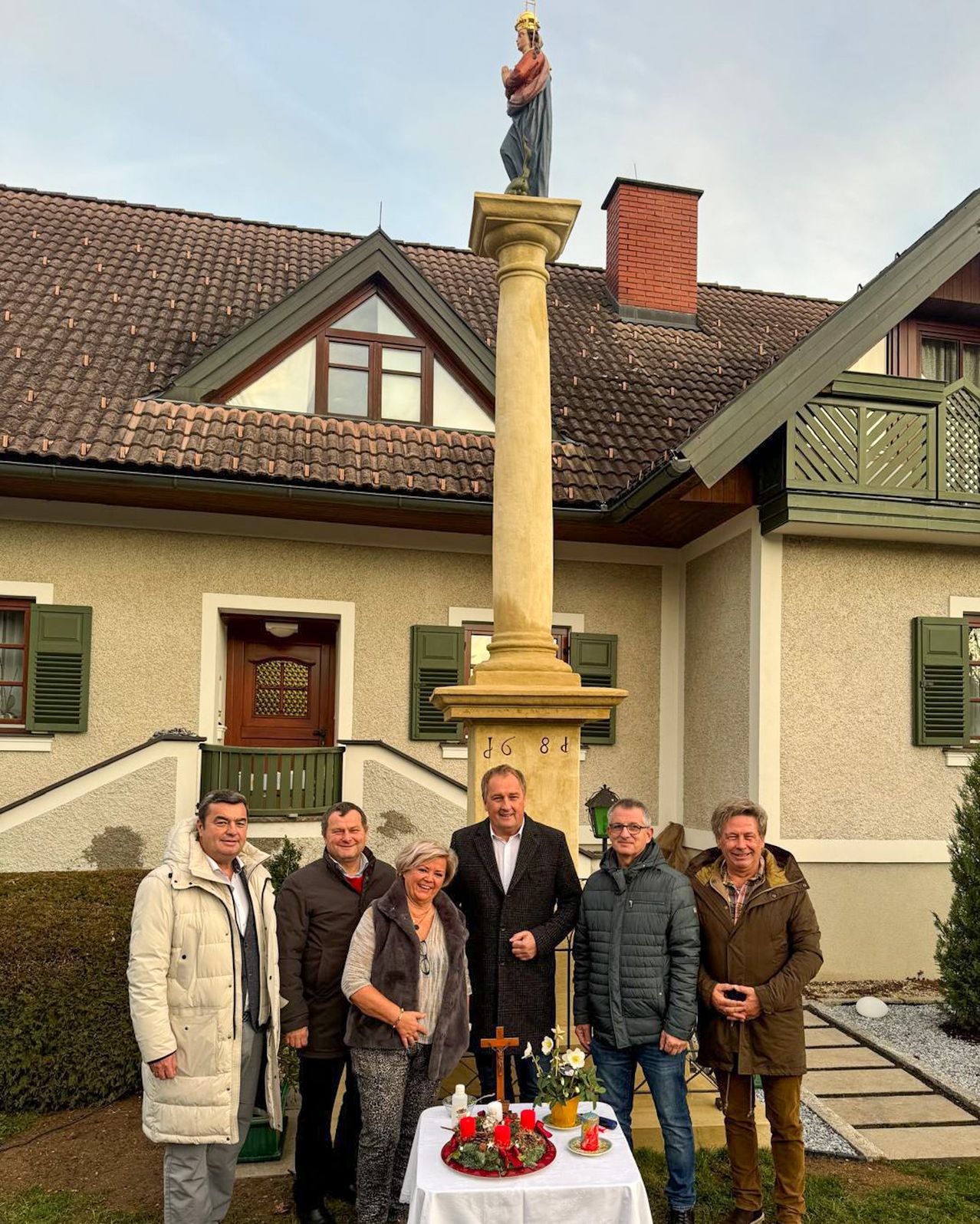 Eine Gruppe von Menschen steht vor einem Haus mit einem hohen Pfeiler. Das Haus hat einen Schornstein, eine Terrasse und grüne Fensterläden. Der Pfeiler hat die Nummer 1684 darauf. Auf dem Boden befindet sich ein Kreuz und Blumen.