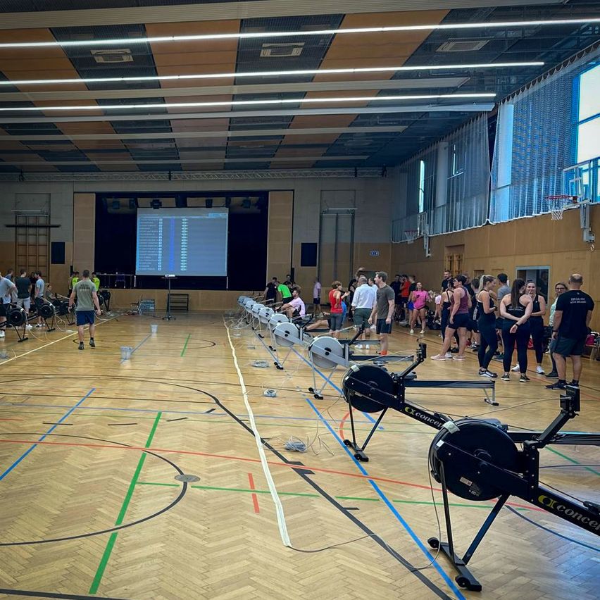 In einer Turnhalle mit Basketballplatz versammeln sich viele Menschen. Sie tragen Sportschuhe und einige benutzen Rudergeräte. An der Wand befindet sich eine Projektionsleinwand.