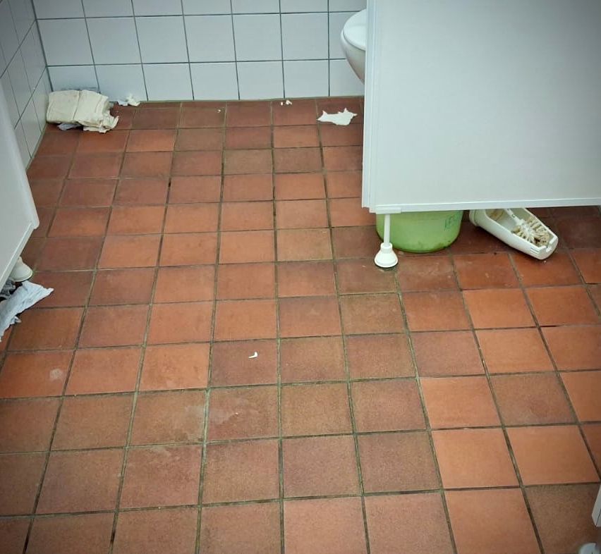 Ein Badezimmerboden mit braunen Fliesen, einer weißen Toilette und einigen Abfällen. Ein grüner Behälter mit einer weißen Bürste steht auf dem Boden.