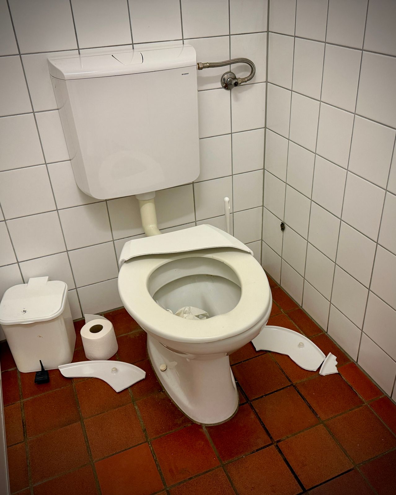 Eine beschädigte weiße Toilette mit zerbrochenem Sitz und Tankdeckel in einem Badezimmer mit weißen Fliesen und einem roten Boden.
