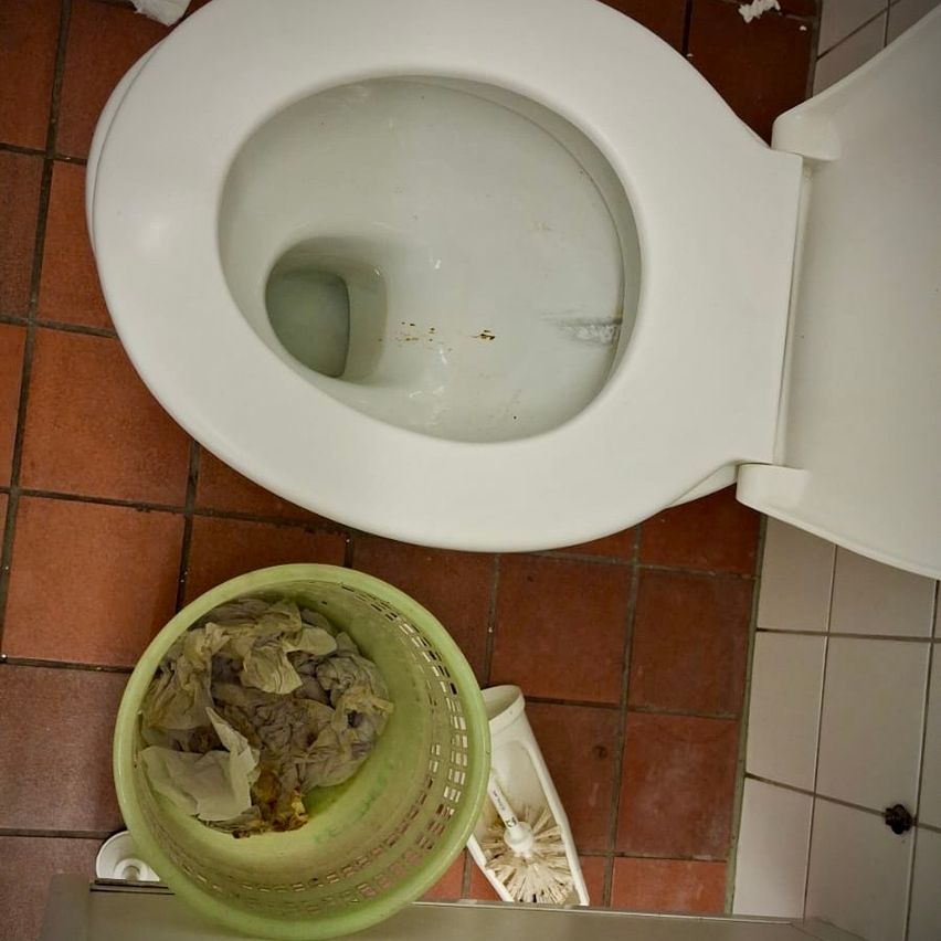 Ein Badezimmer mit einer weißen Toilettenschüssel, die Rückstände zeigt, und einem grünen Korb mit benutzten Taschentüchern daneben. Der Boden ist gefliest.