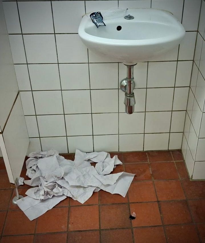 Ein weißer Waschbecken ist an einer gefliesten Wand montiert, mit einem Haufen zerknitterter Papiere auf dem Boden. Ein Stück Papier ist am Wasserhahn befestigt.