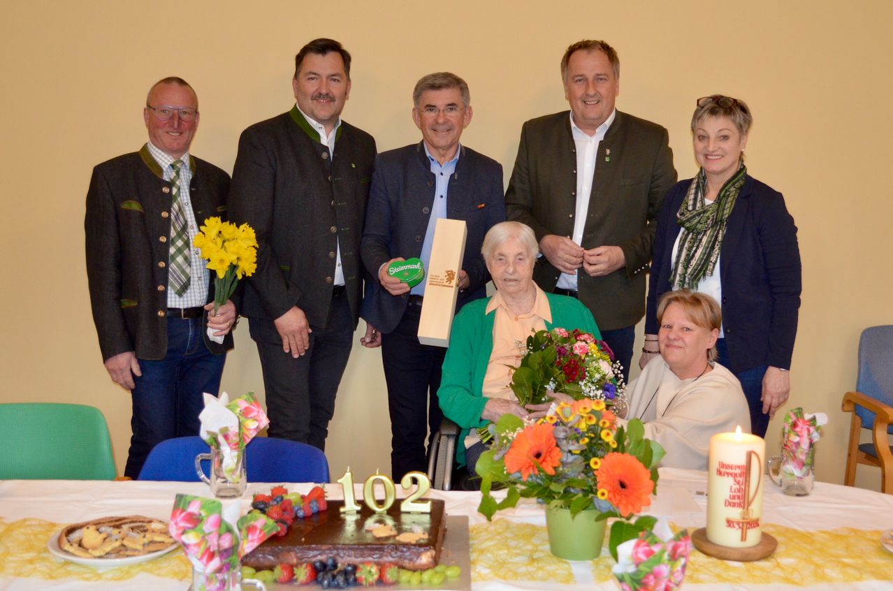 A group of people are gathered around a table celebrating a birthday. A woman is in a wheelchair receiving flowers. A cake with the number 102 is present. The people are dressed in formal attire.
