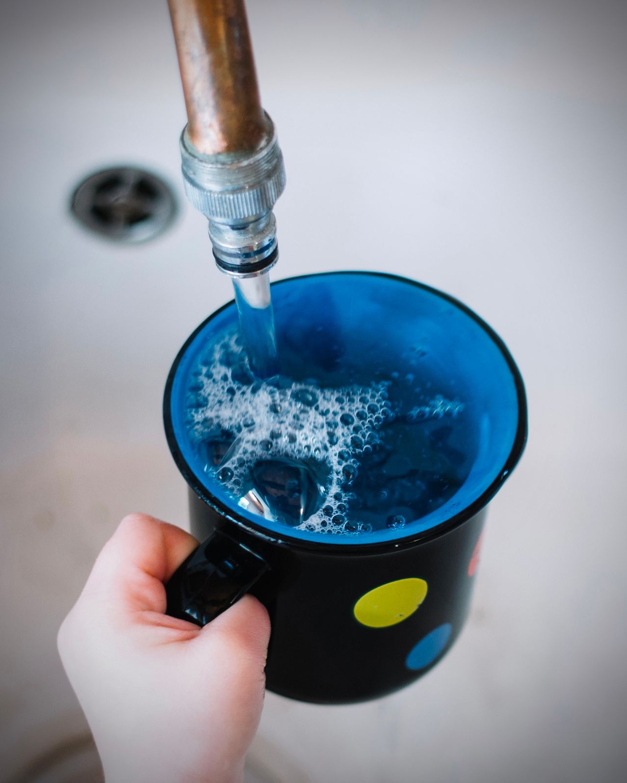 A person fills a blue mug with water from a copper pipe, with bubbles visible in the water.