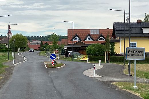 Eine leere Straße in St. Peter am Ottersbach mit Häusern, einem blauen Schild und Straßenlaternen.