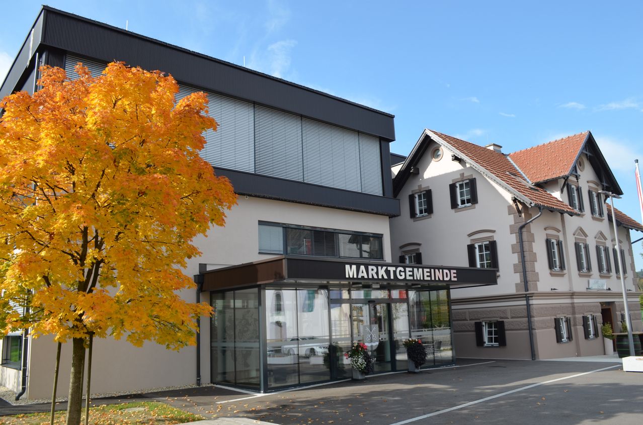 Ein modernes Gebäude mit der Aufschrift 'Marktgemeinde' über dem Eingang. Das Gebäude hat Glaswände und einen braunen Vorbau. Ein Baum mit gelben Blättern steht links.