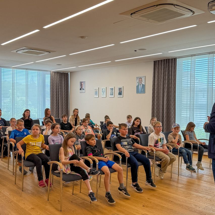 A room with students and adults seated in chairs, looking at a person standing. The room has wooden floors and white walls with picture frames.