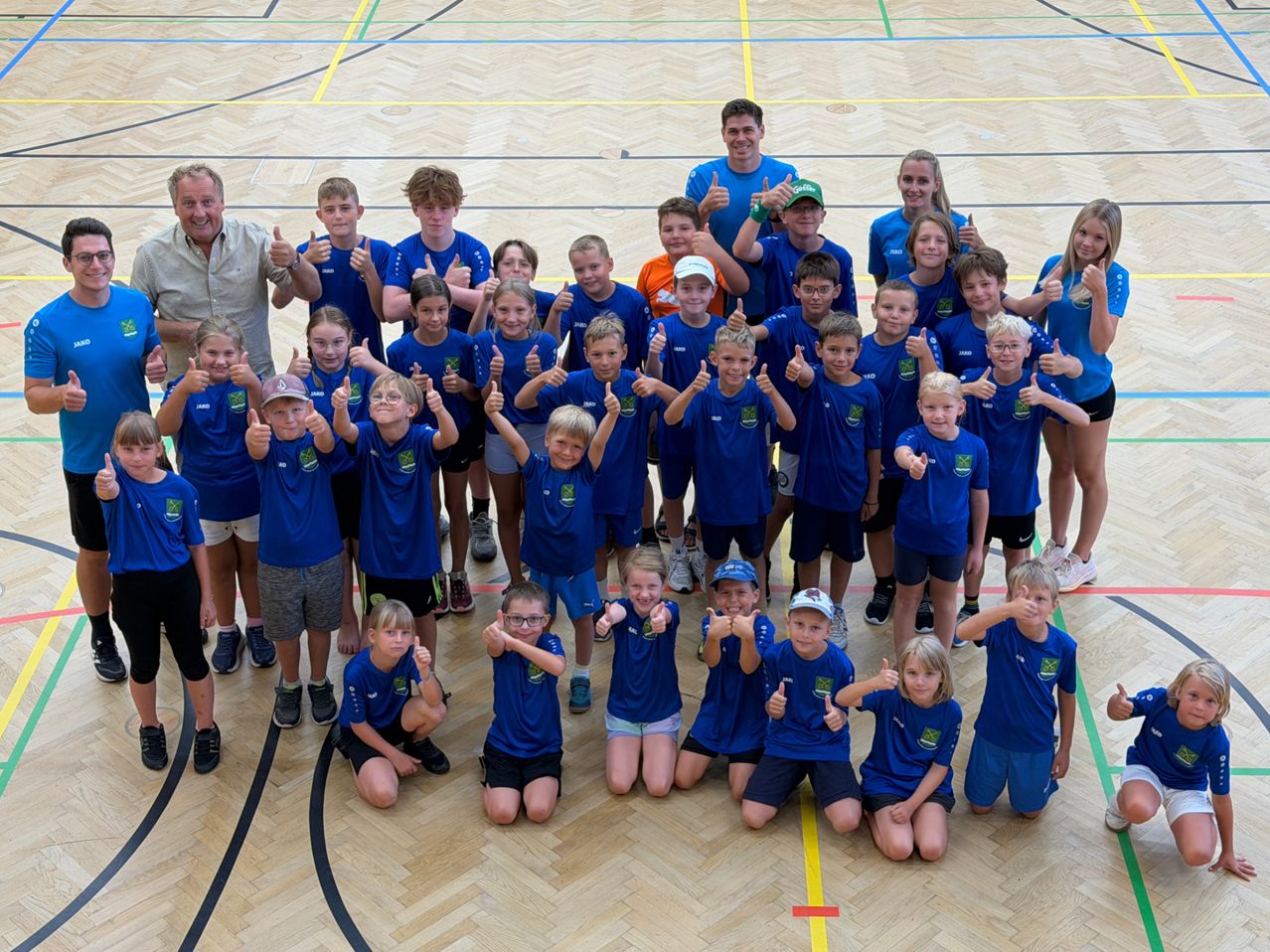 Eine Gruppe von Kindern und Erwachsenen in blauen Shirts und Shorts posiert für ein Foto in einer Turnhalle. Sie alle zeigen Daumen hoch.