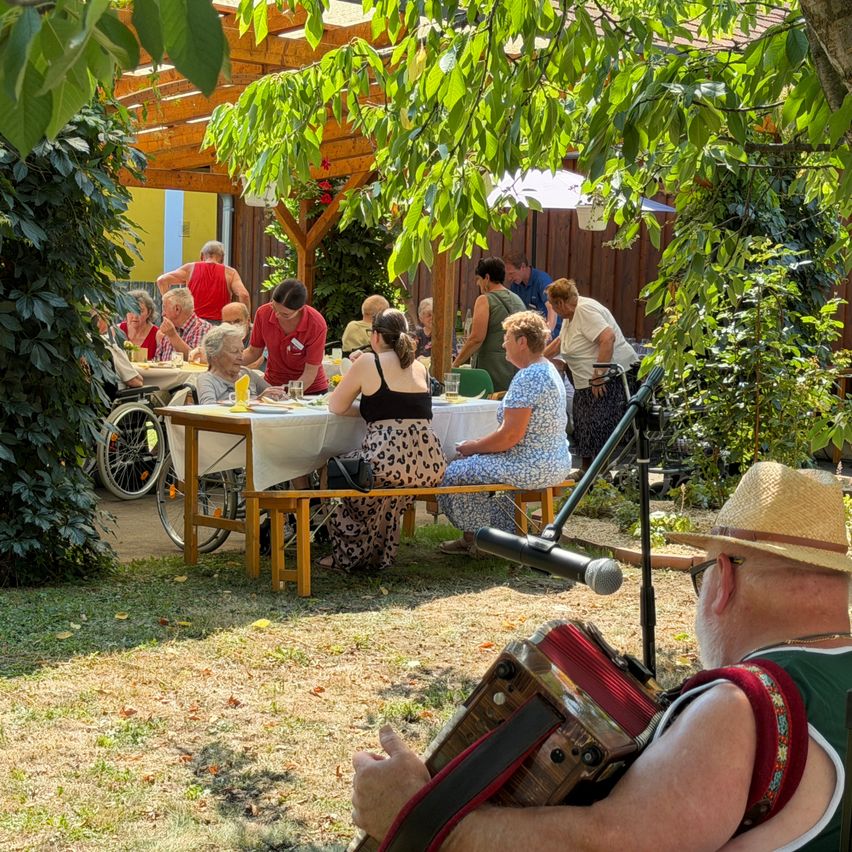 Eine Gruppe älterer Menschen sitzt an Tischen in einem Garten, einige essen, während andere einem Mann beim Akkordeonspielen zuhören.