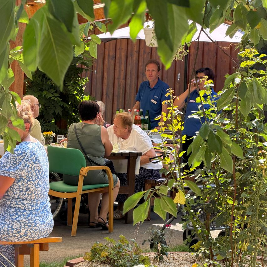 Eine Gruppe von Menschen sitzt um einen Tisch in einem Garten, ein Mann steht und eine Frau trinkt aus einem Glas.