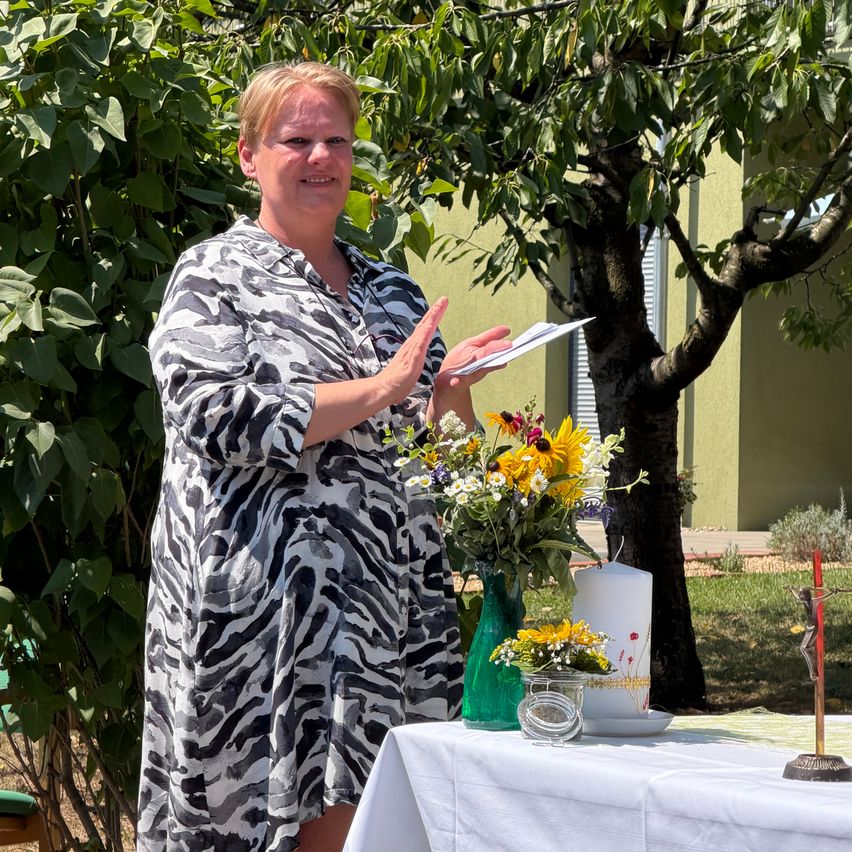 Eine Frau in einem Zebra-Druckkleid steht im Freien und klatscht in die Hände, während sie ein Buch in der Hand hält. Vor ihr steht ein Tisch mit Blumen, einer Kerze und einem Kruzifix. Hinter ihr sind ein Baum und ein Gebäude zu sehen.