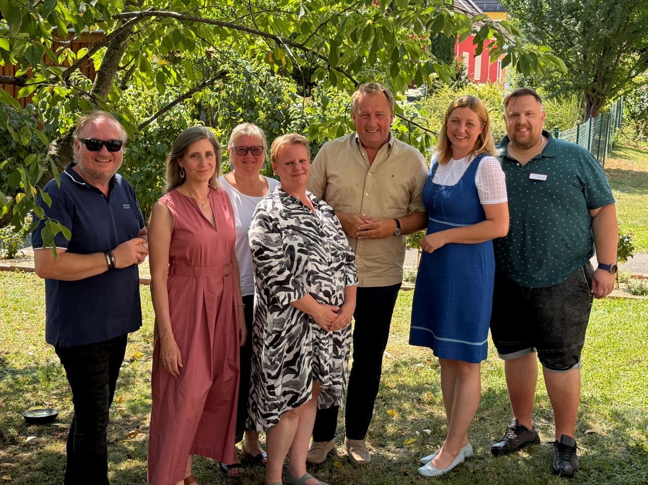 Eine Gruppe von sieben Personen, die lächelnd und posierend für ein Foto in einem Garten mit Bäumen und einem roten Gebäude im Hintergrund.