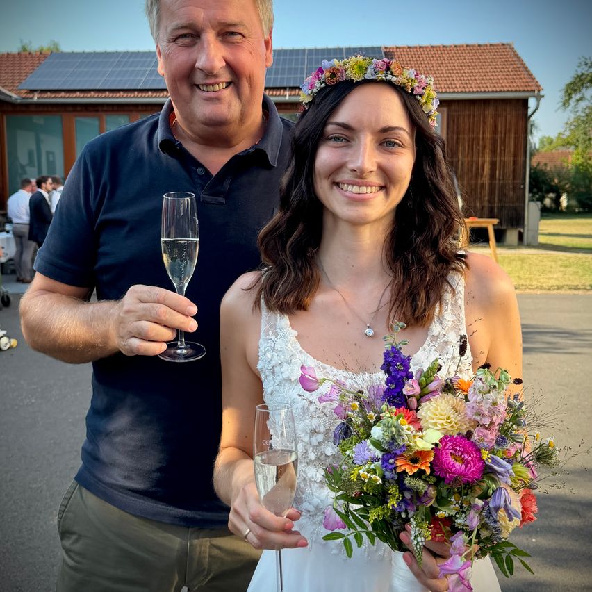 Ein Mann und eine Frau, wahrscheinlich ein Paar, stehen draußen. Der Mann hält ein Glas Champagner und die Frau eine Blumenbouquet. Sie lächeln beide.