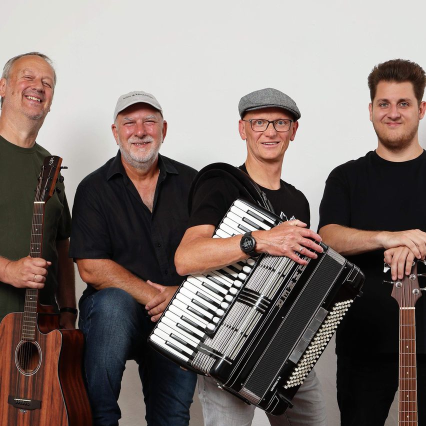 Vier Männer posieren für ein Foto, jeder hält ein Musikinstrument. Der Mann auf der linken Seite hält eine Gitarre, der zweite Mann von links hält ein Akkordeon, der dritte Mann hält eine Gitarre und der Mann auf der rechten Seite hält eine Gitarre.