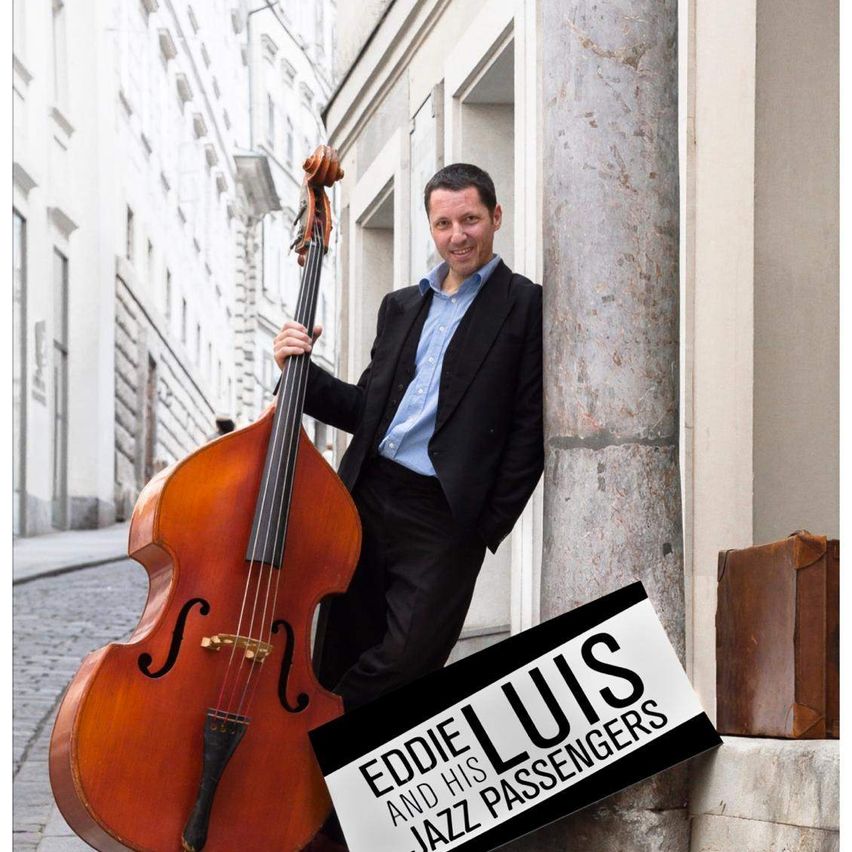 Eddie Luis, ein Musiker, steht neben einem großen Kontrabass, lächelt und lehnt sich gegen eine Steinsäule. Der Hintergrund zeigt eine Kopfsteinpflasterstraße und Gebäude.