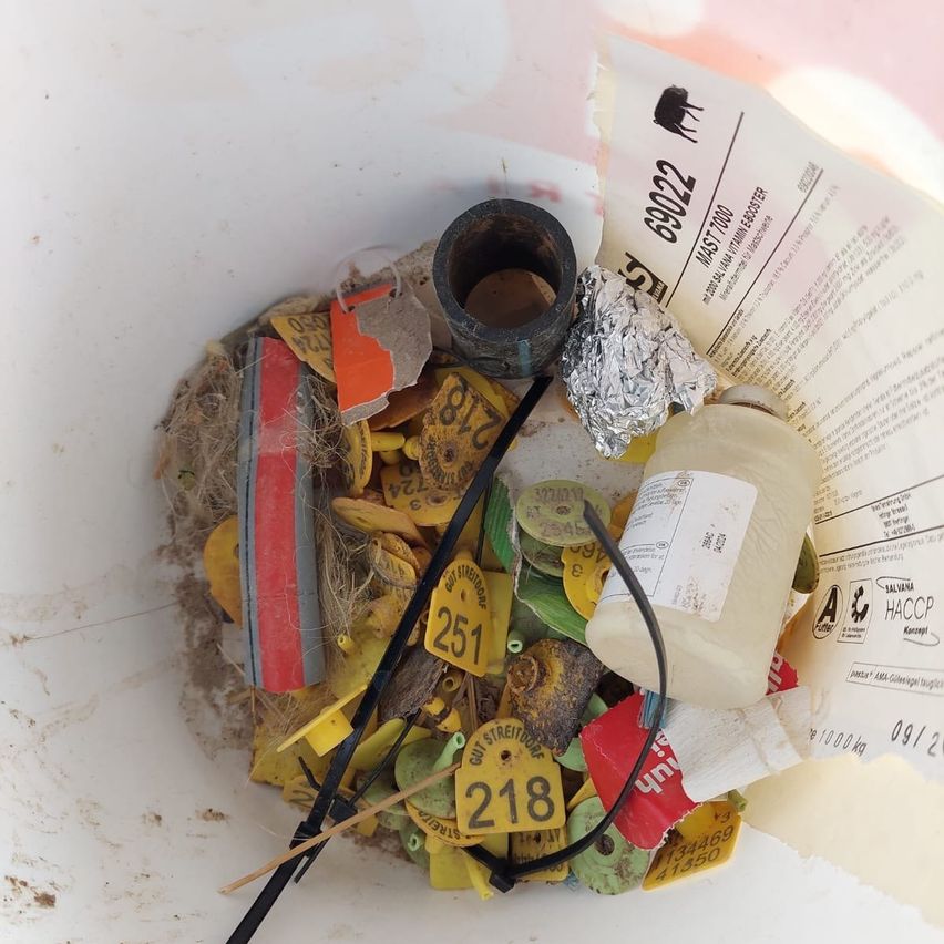Ein weißer Eimer enthält verschiedene Gegenstände, darunter gelbe Etiketten mit Nummern, ein Klebeband, einen folienbedeckten Gegenstand, eine Plastikflasche und Papierstücke.