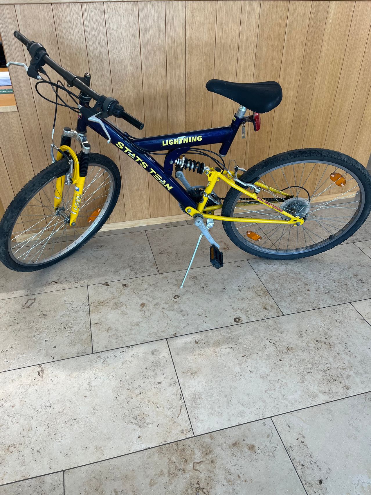 A blue and yellow bicycle with 'Lightning' and 'Stats Team' printed on it is parked on a tiled floor in front of a wooden wall.