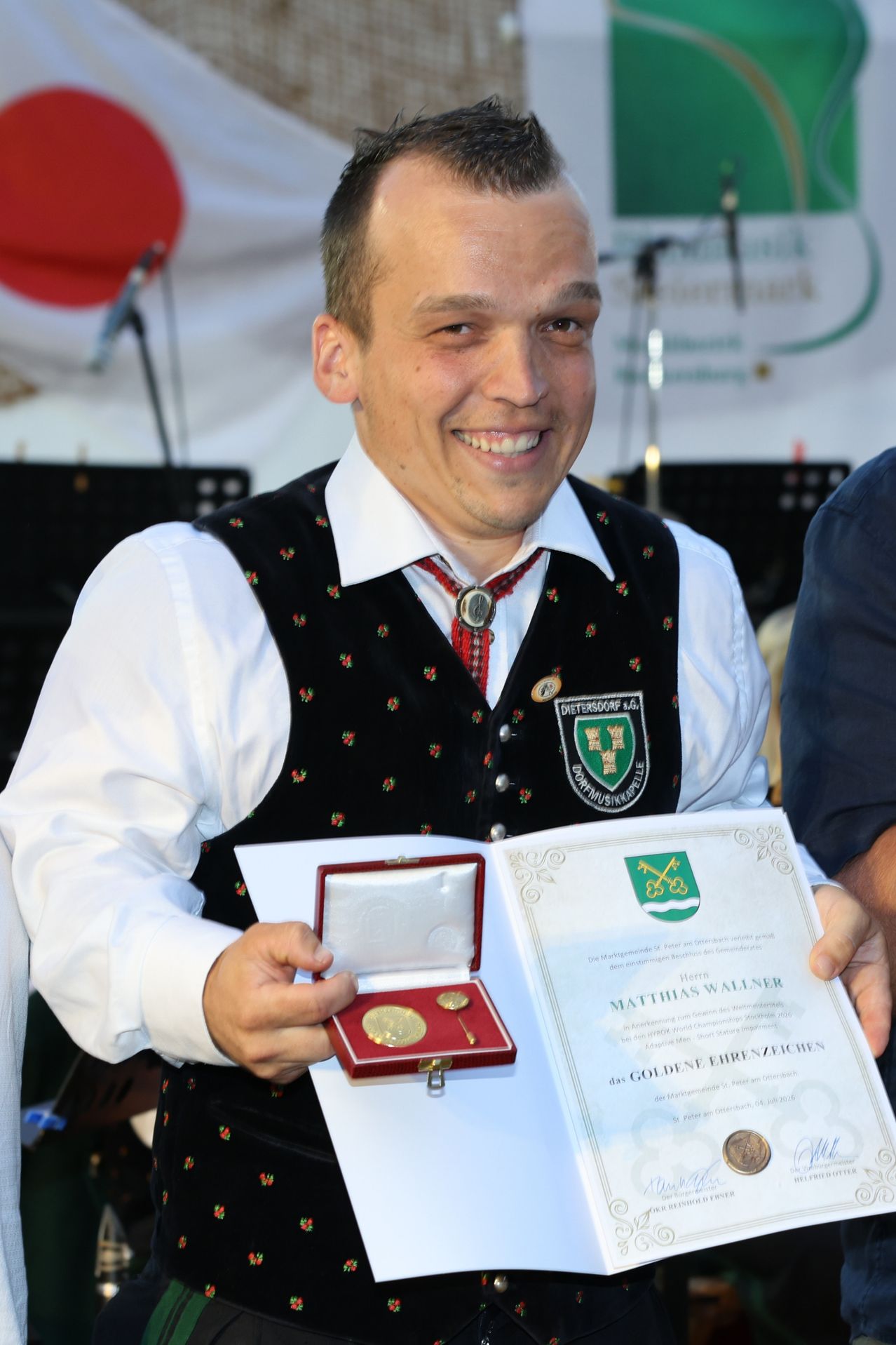 A man smiles as he holds a golden medal and a certificate. He wears a traditional black vest with red flowers. Behind him, someone else holds a red box containing more medals.