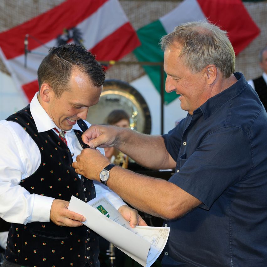 A man in a blue shirt is pinning a medal onto another man's vest. Both are smiling. They are in front of a flag and a drum.