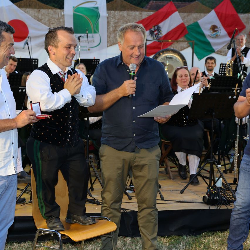 A man is giving an award to another man standing on a chair on a stage. Behind them is an orchestra and a man holding a microphone.