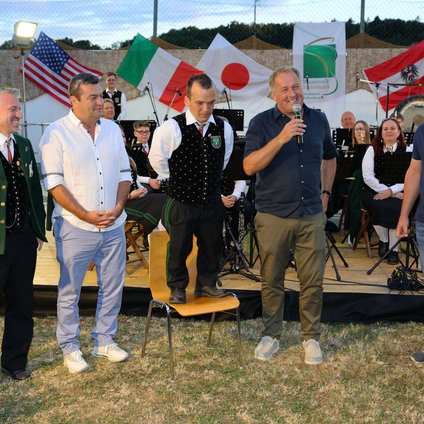 A man holds a microphone, smiling, while standing on a chair. He is surrounded by four others on a stage, with musical instruments and flags in the background.
