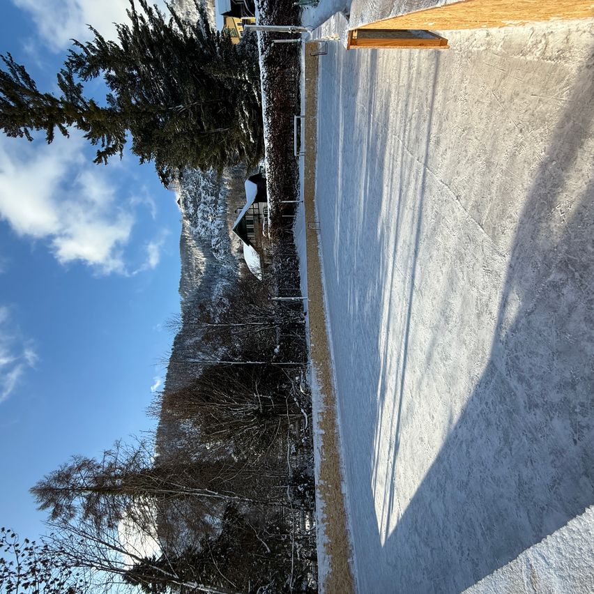 Ein Berghang mit Bäumen, einem Gebäude und einer vereisten Oberfläche unter einem blauen Himmel mit Wolken.