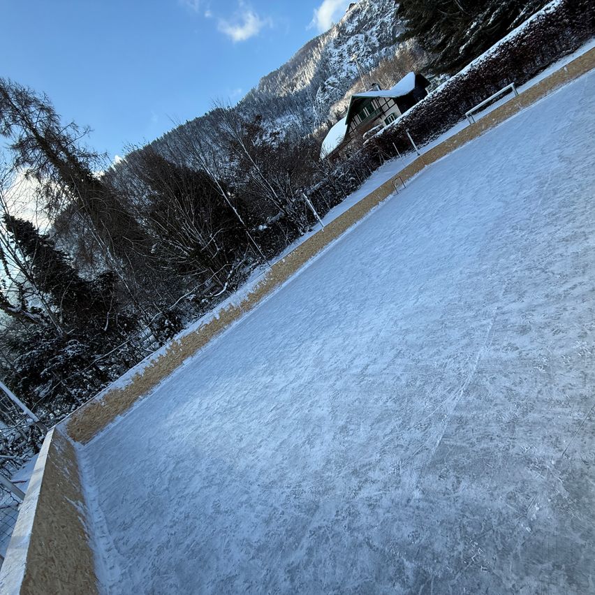 Eine leere Eisbahn, die mit Schnee bedeckt ist, umgeben von schneebedeckten Bäumen und Bergen. Im Hintergrund ist ein Haus mit Schnee auf dem Dach zu sehen.