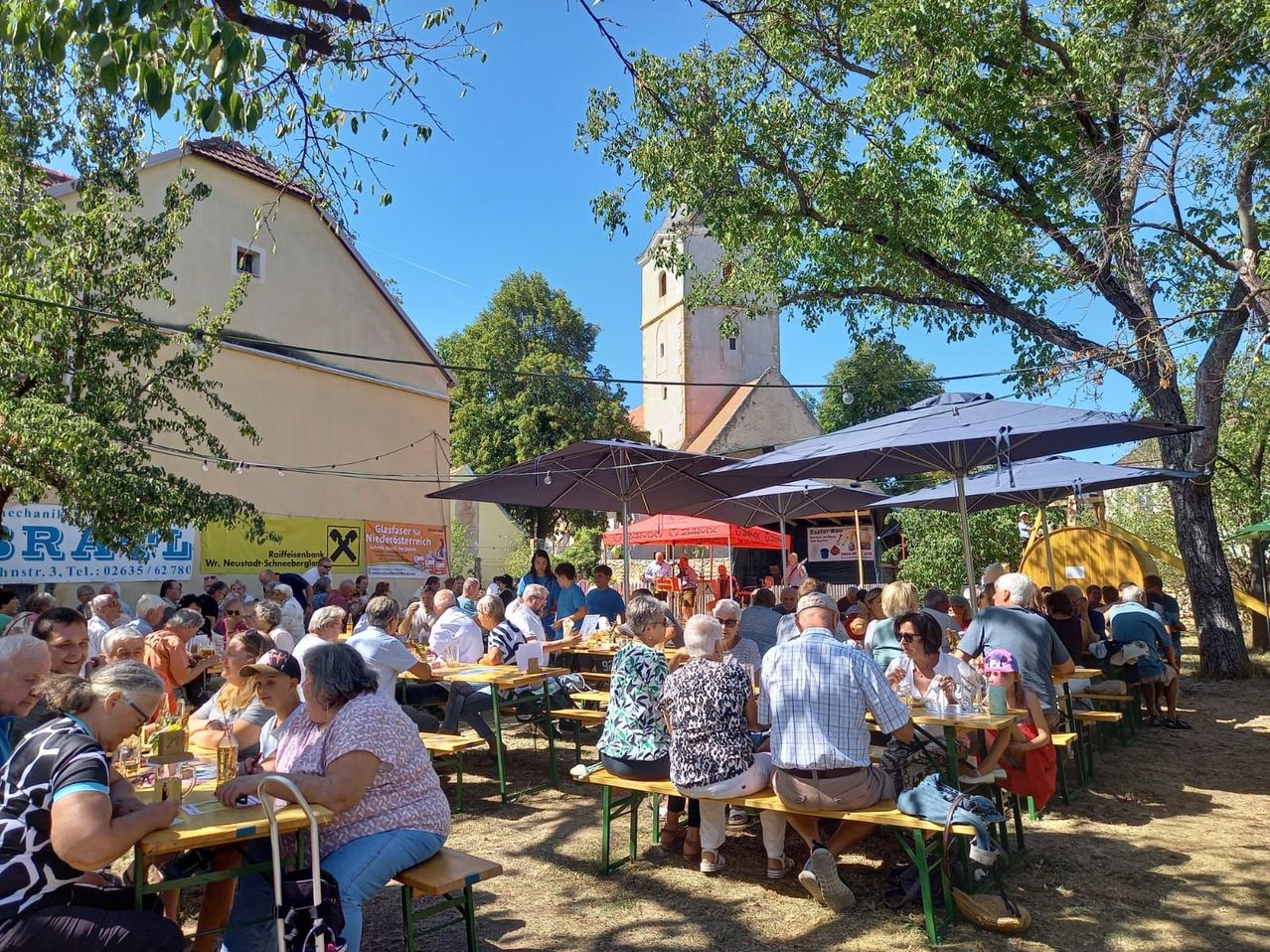 A large group of people are gathered in an outdoor area, possibly for a festival. They are seated around tables under umbrellas, with a building and a tower in the background.