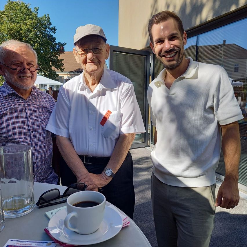 Three men stand outside, smiling, with one holding a cup of coffee. The man in the middle wears a hat and a watch. The background shows a building and a tree.