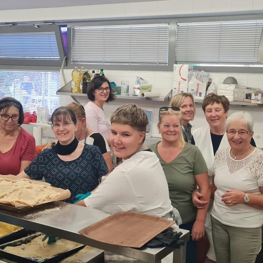 A group of women in a kitchen, smiling and posing for a photo. One woman is wearing gloves and holding a tray with food. Behind them are shelves with bottles, boxes, and other items.