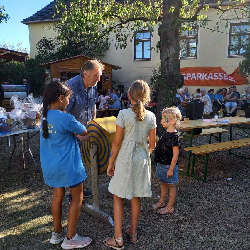 An older man is talking to two young girls in front of a yellow spinning wheel at an outdoor event. Behind them, there are tables with items and a crowd of people sitting on benches.